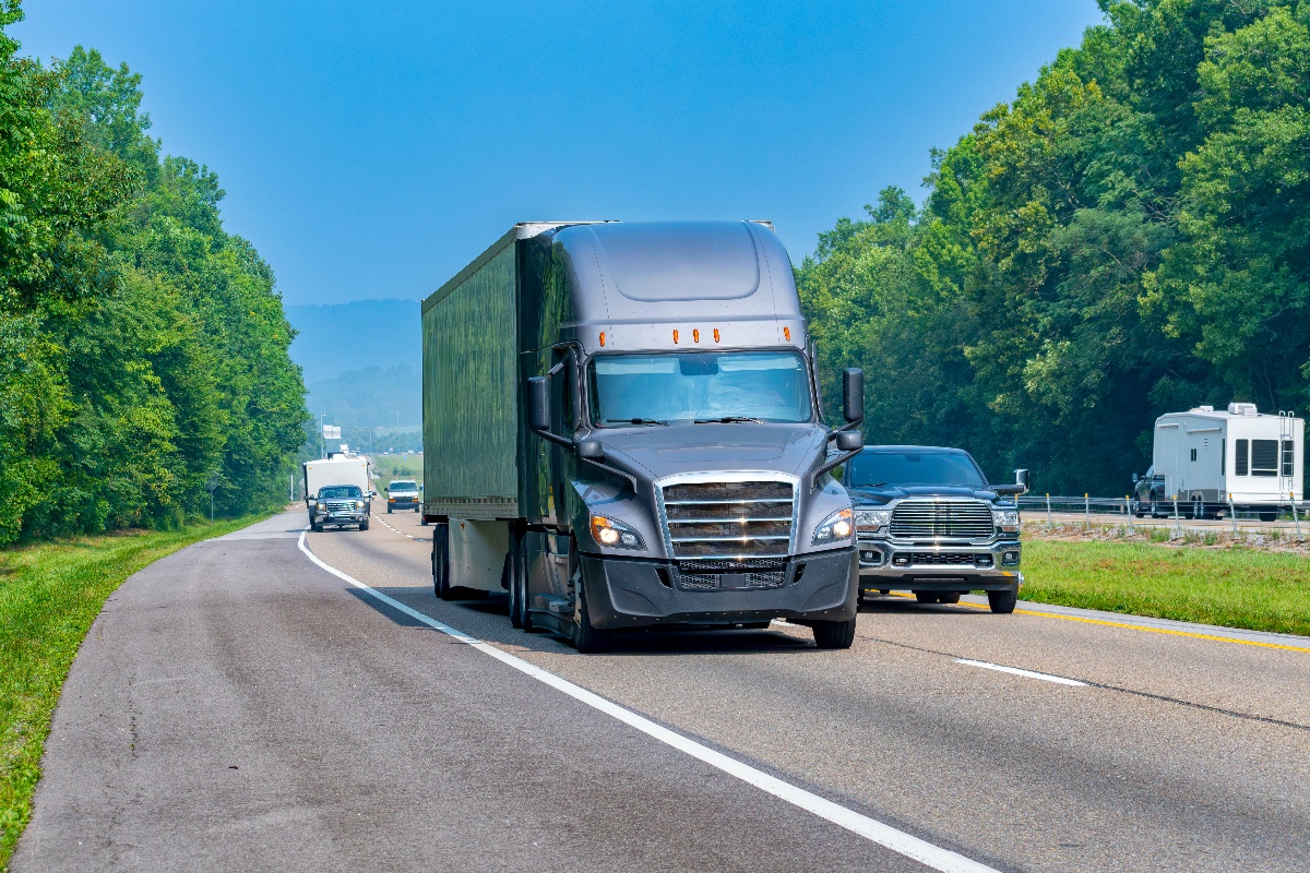 A grey semi-truck drives down a busy highway surrounded by trees. 