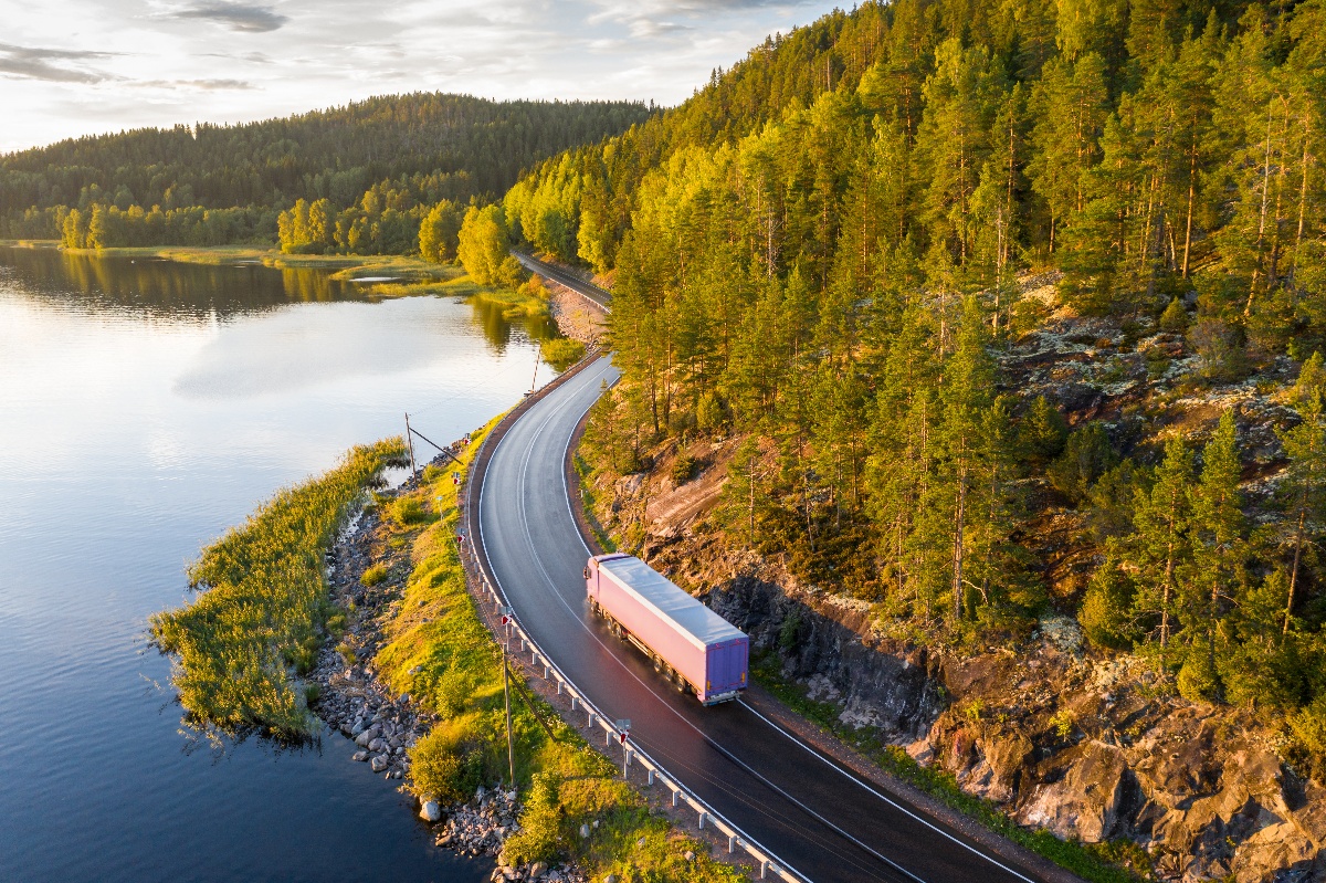A semi-truck drives around a corner of a mountain road across from a lake.