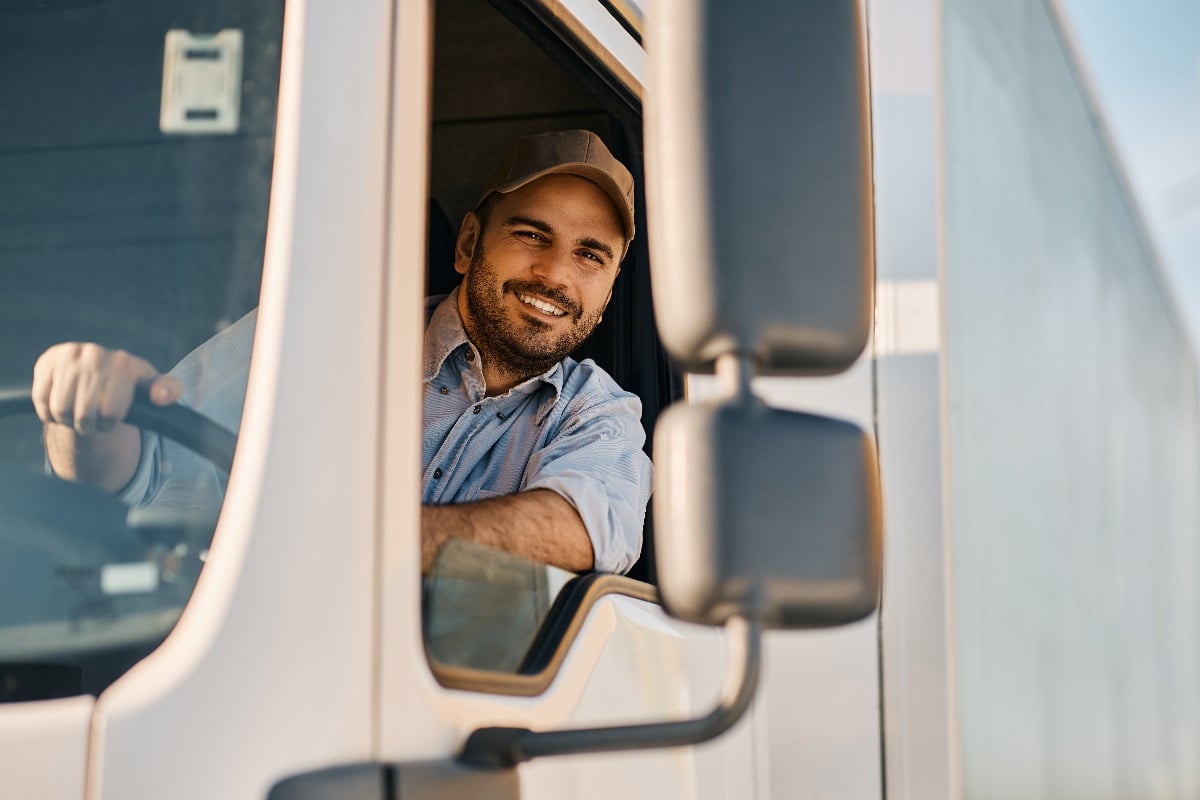 A male semi truck driver smiles as he drives a loaded semi-truck.