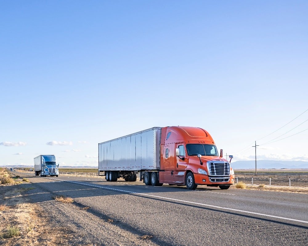 An orange semi truck drives ahead of a blue semi-truck on a highway. 